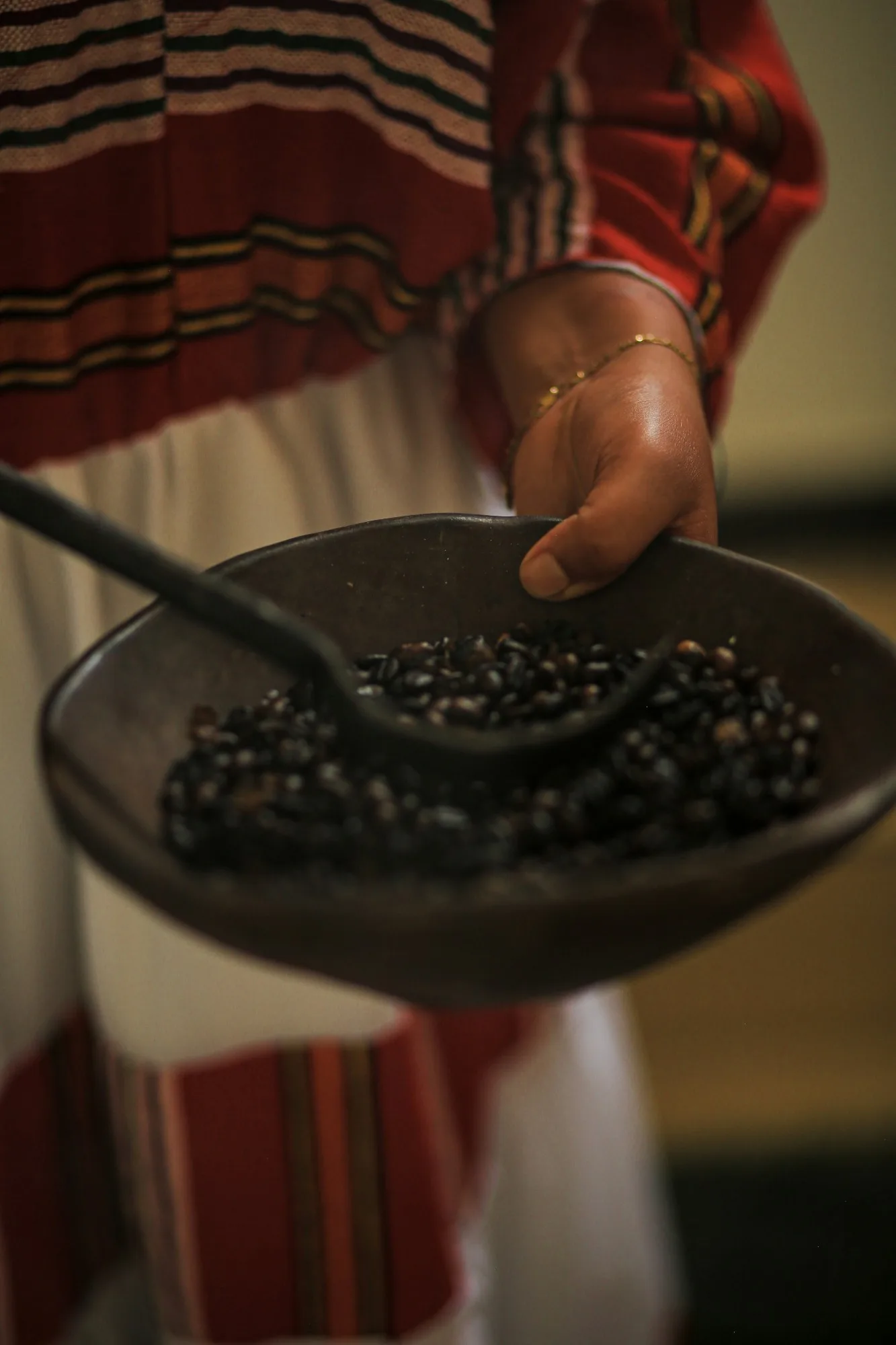 A hand holding a clay pan of freshly roasted coffee beans, with a traditional Ethiopian woven garment behind